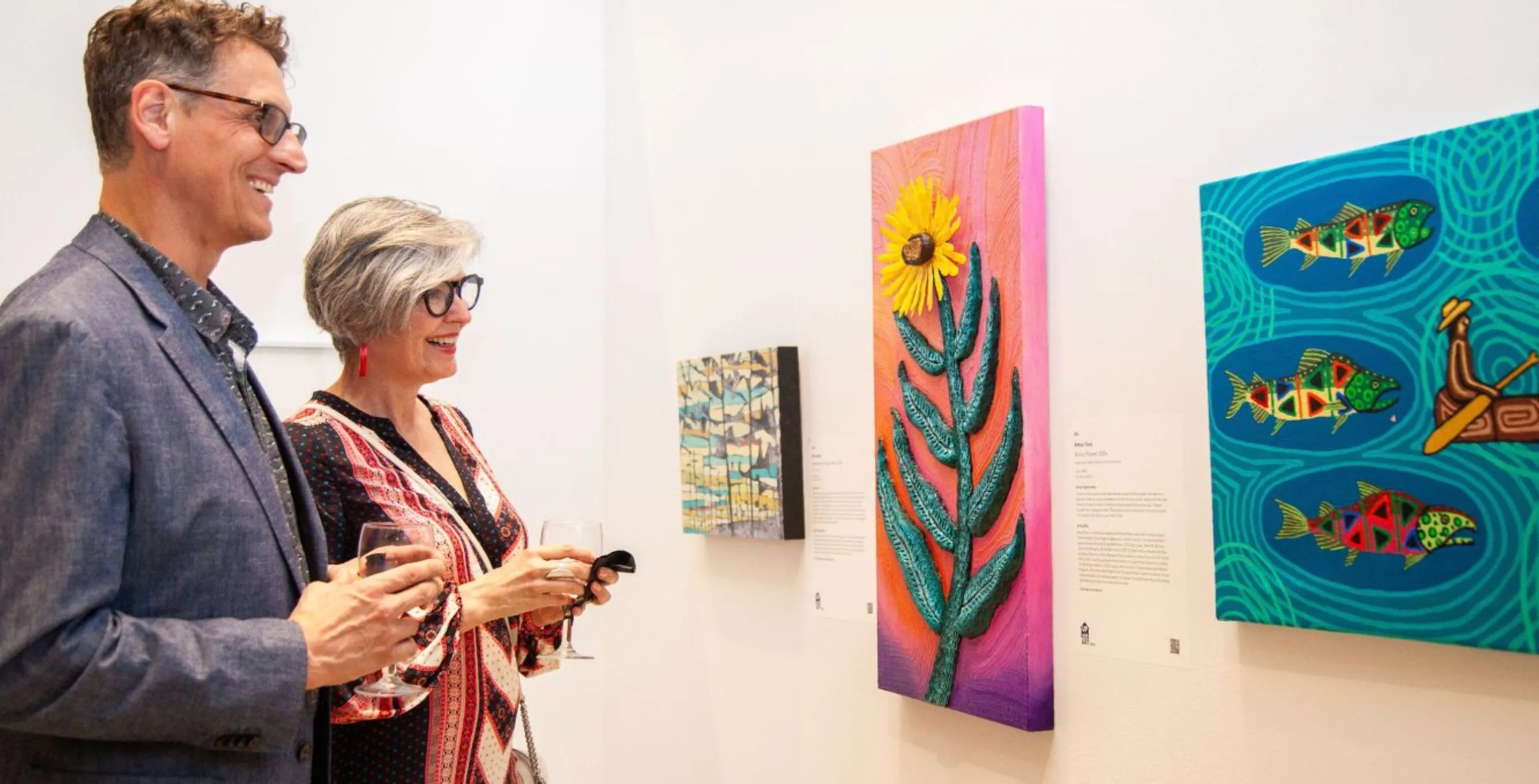 Two people are standing in front of three vibrant works of art, admiring them and conversing.
