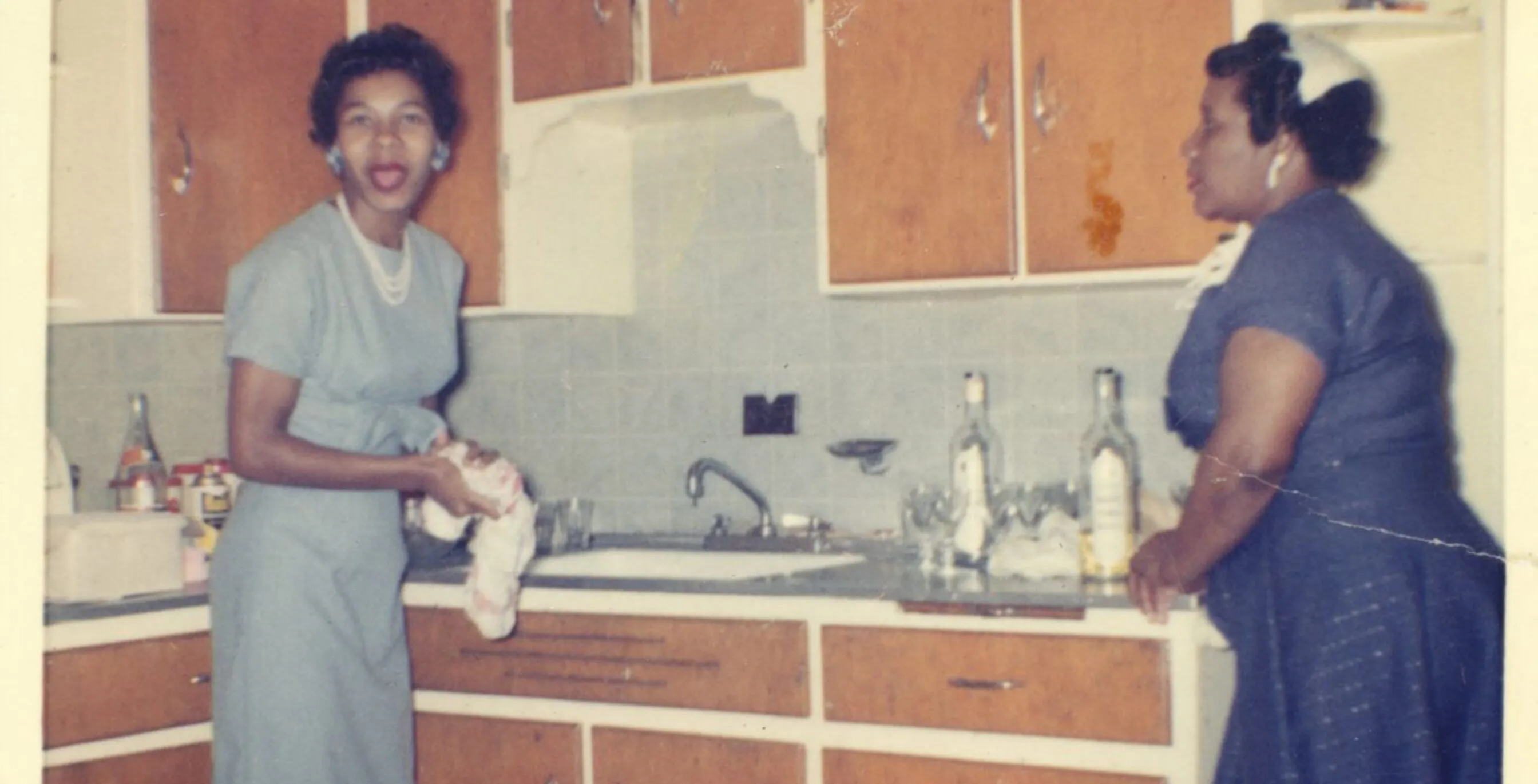 two women in formal attire stand in a 1960s kitchen with wood cupboards and tile backsplash. The woman on the left is looking at the camera.