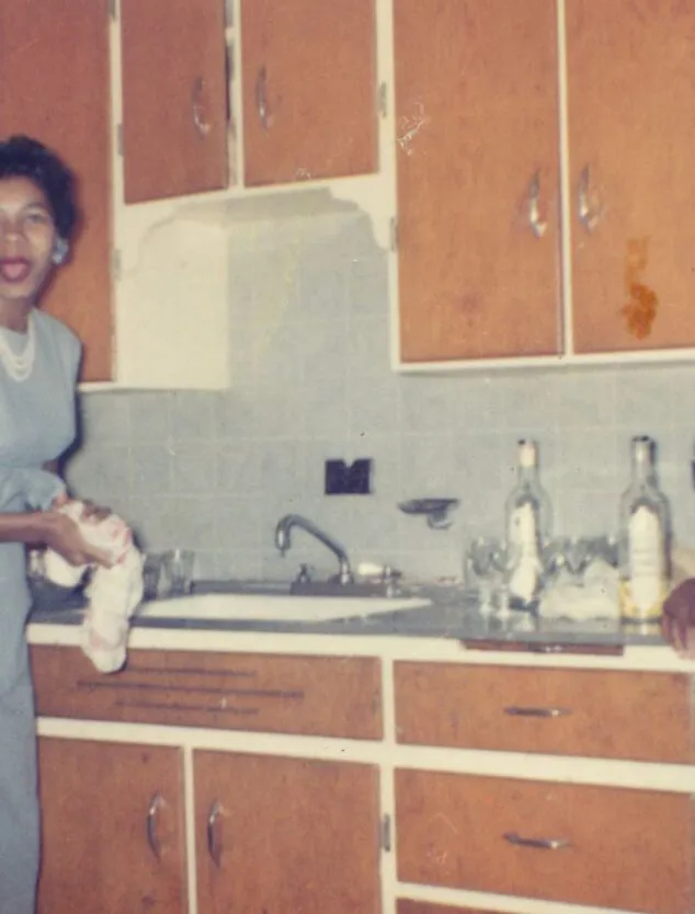 two women in formal attire stand in a 1960s kitchen with wood cupboards and tile backsplash. The woman on the left is looking at the camera.