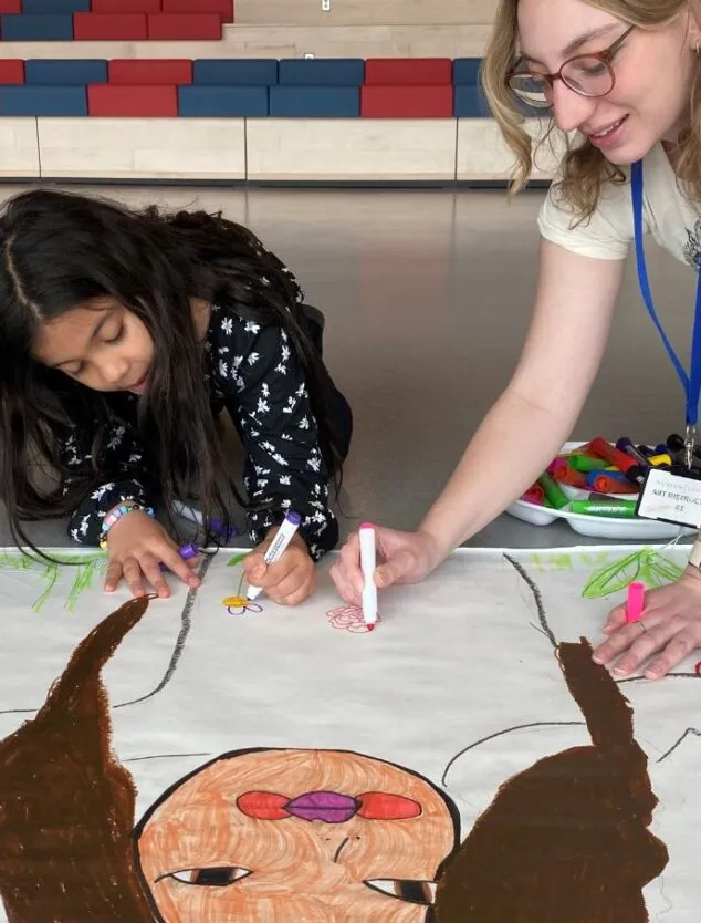a school-aged child draws a larger than life character alongside an art instructor